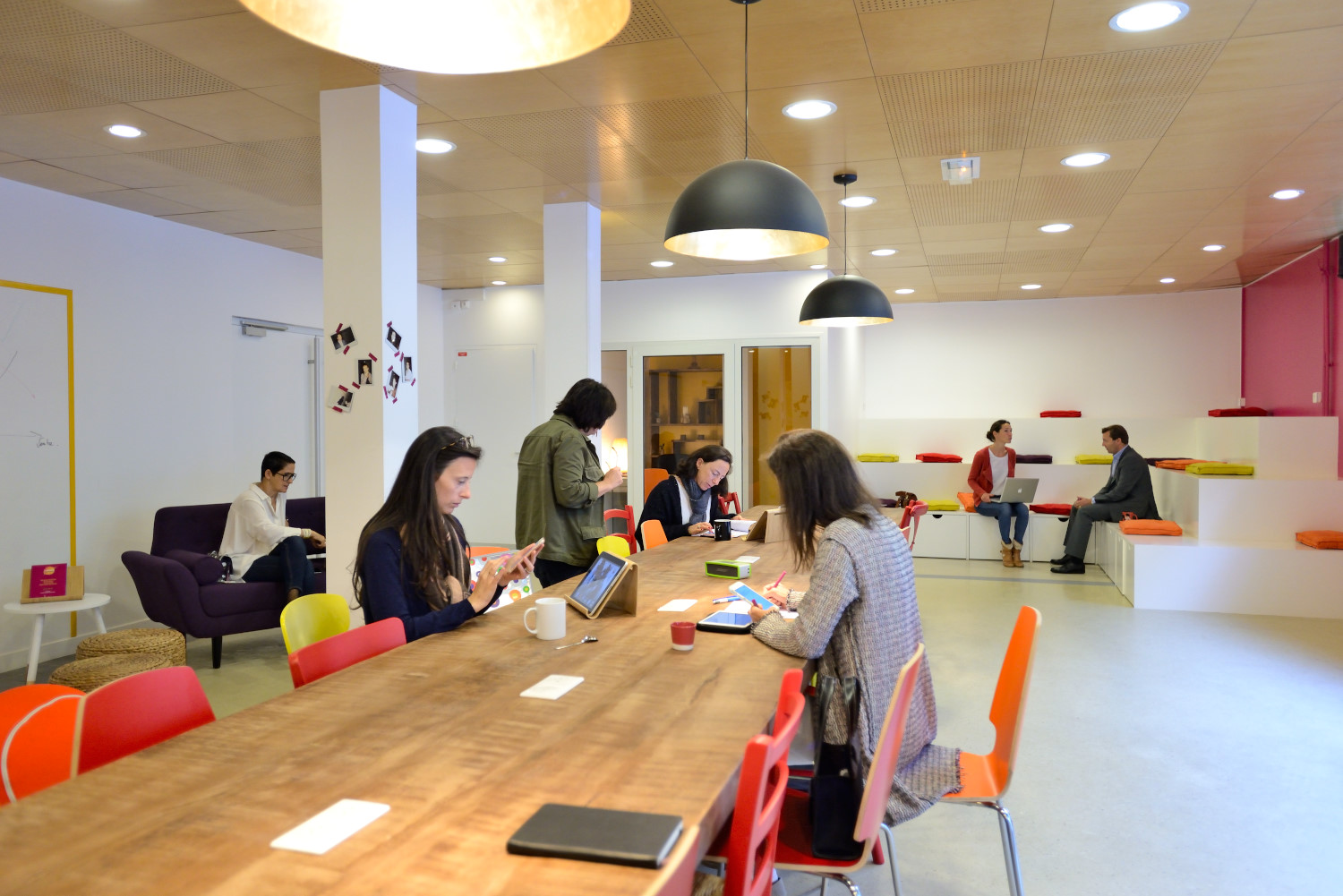 Un espace de bureau moderne avec des personnes travaillant à une longue table en bois commune et d'autres utilisant des sièges à gradins le long des murs. La pièce comprend des chaises colorées, des luminaires suspendus et une atmosphère détendue et collaborative parfaite pour une séance photo portrait pour CV LinkedIn et site web.