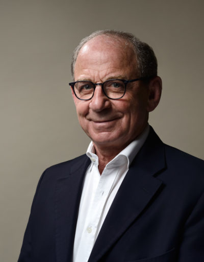 Une séance photo portrait pour CV, LinkedIn et site web capture un homme aux cheveux gris courts et lunettes, vêtu d'un blazer sombre sur une chemise blanche, souriant légèrement devant un fond uni couleur ivoire.