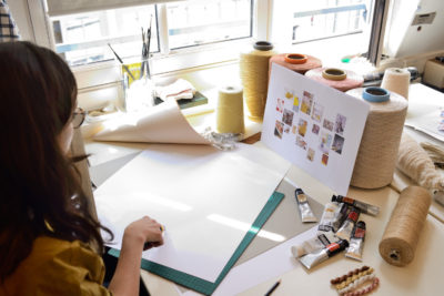 Une personne aux cheveux longs et portant des lunettes est vue de dos, assise à un bureau rempli de fournitures artistiques comme des tubes de peinture et de grandes bobines de fil. À côté d'eux se trouve une feuille de papier vierge à côté d'un collage de photos épinglé sur un tableau, se préparant peut-être à une séance photo portrait pour CV linkedin et site web. La pièce dispose de grandes fenêtres laissant entrer la lumière naturelle.