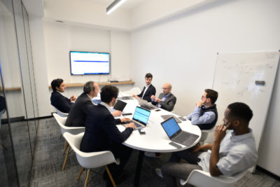 Un groupe de sept personnes, habillées en tenue professionnelle, sont assises autour d'une table ronde blanche dans une salle de conférence moderne, engagées dans une discussion. Des ordinateurs portables sont ouverts devant la plupart des participants, avec un grand écran et un tableau blanc visibles en arrière-plan, parfait pour une séance photo portrait pour CV LinkedIn et site web.