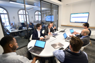 Un groupe de six hommes assis autour d'une table dans une salle de conférence moderne, chacun avec un ordinateur portable ouvert devant eux. Un homme est debout et fait une présentation tandis que d'autres écoutent et prennent des notes. La salle a des murs en verre, révélant un autre espace de travail où se déroule une séance photo portrait pour CV LinkedIn et site web.