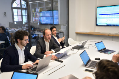 Quatre hommes en tenue professionnelle sont assis autour d'une table avec des ordinateurs portables dans un bureau moderne. Un grand écran sur le mur affiche une feuille de calcul. Des étagères avec des livres et des documents sont visibles en arrière-plan. Un homme parle pendant que d'autres écoutent et tapent sur leurs ordinateurs portables, créant une scène idéale pour une séance photo portrait pour CV LinkedIn et site web.