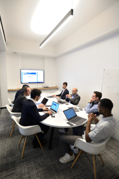 Un groupe diversifié de sept personnes est assis autour d'une table blanche dans une salle de réunion de bureau moderne, avec des ordinateurs portables ouverts et des papiers étalés. Un grand écran sur le mur affiche une présentation et un tableau blanc est rempli de notes. Lumineuse, la salle capture une toile de fond idéale pour une séance photo portrait pour CV LinkedIn et site web.