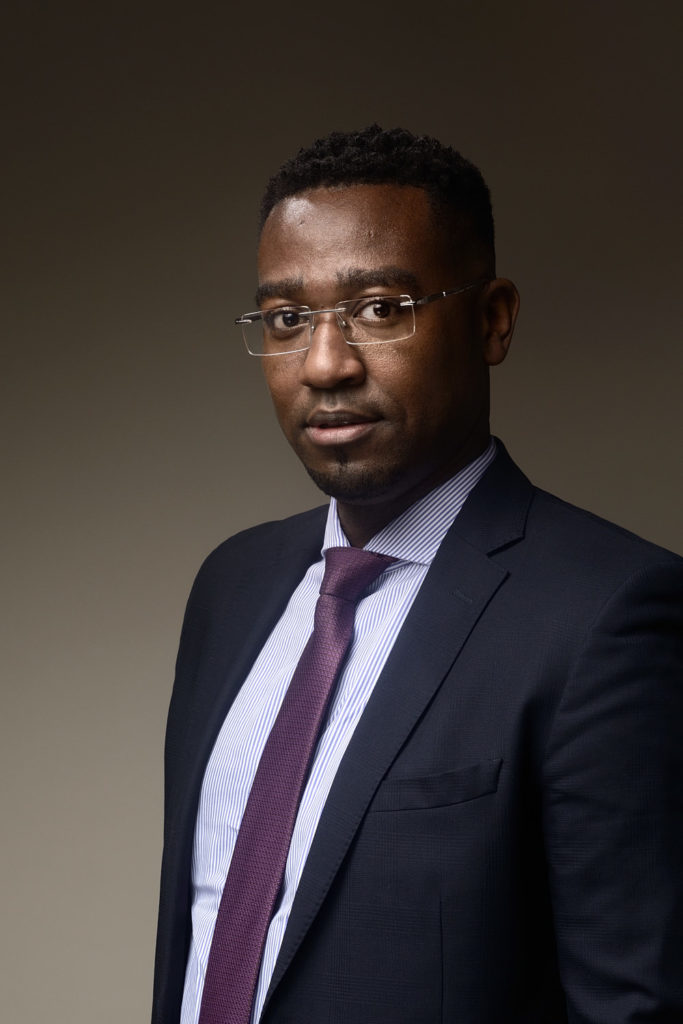 Un homme portant des lunettes, vêtu d'un costume sombre avec une chemise rayée et une cravate marron, se tient debout sur un fond sombre uni. Il a les cheveux courts et une expression calme et confiante, parfaite pour une séance photo portrait pour CV LinkedIn et site web.