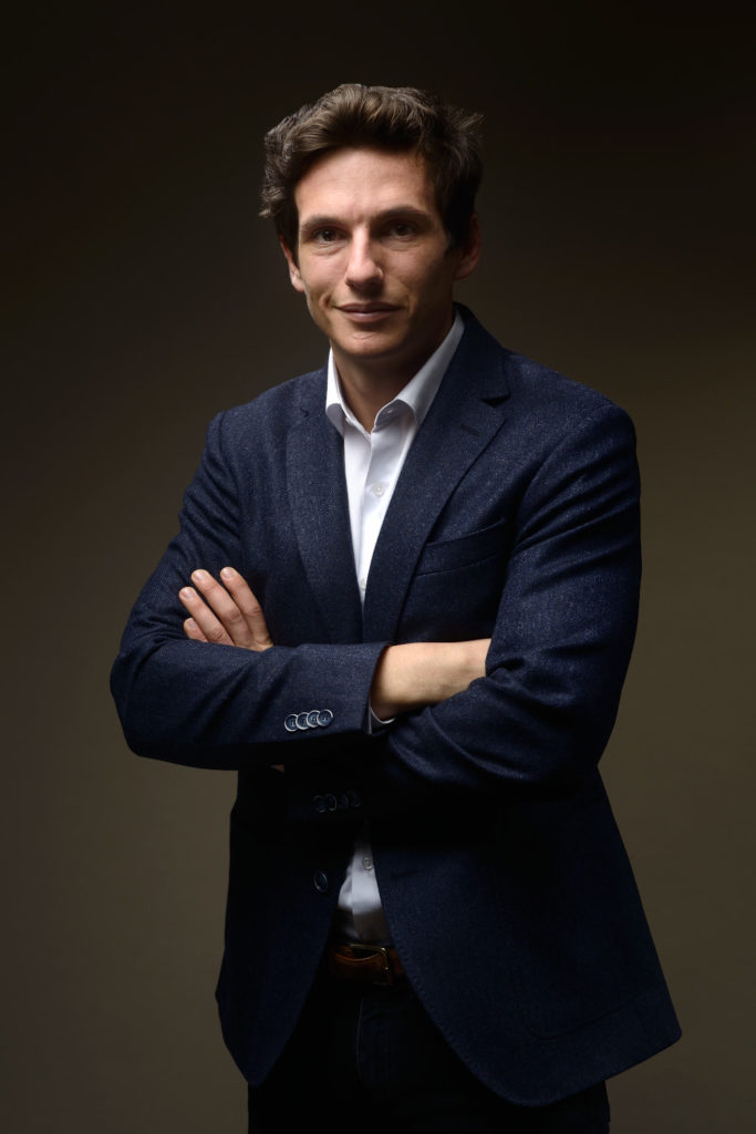 Un homme vêtu d'un blazer bleu foncé se tient debout, les bras croisés, sur un fond sombre. Il a les cheveux courts et foncés et porte une chemise blanche boutonnée. L'éclairage met en valeur son visage et le haut de son corps, créant une apparence professionnelle et confiante, parfaite pour une séance photo portrait pour CV Linkedin et site web.