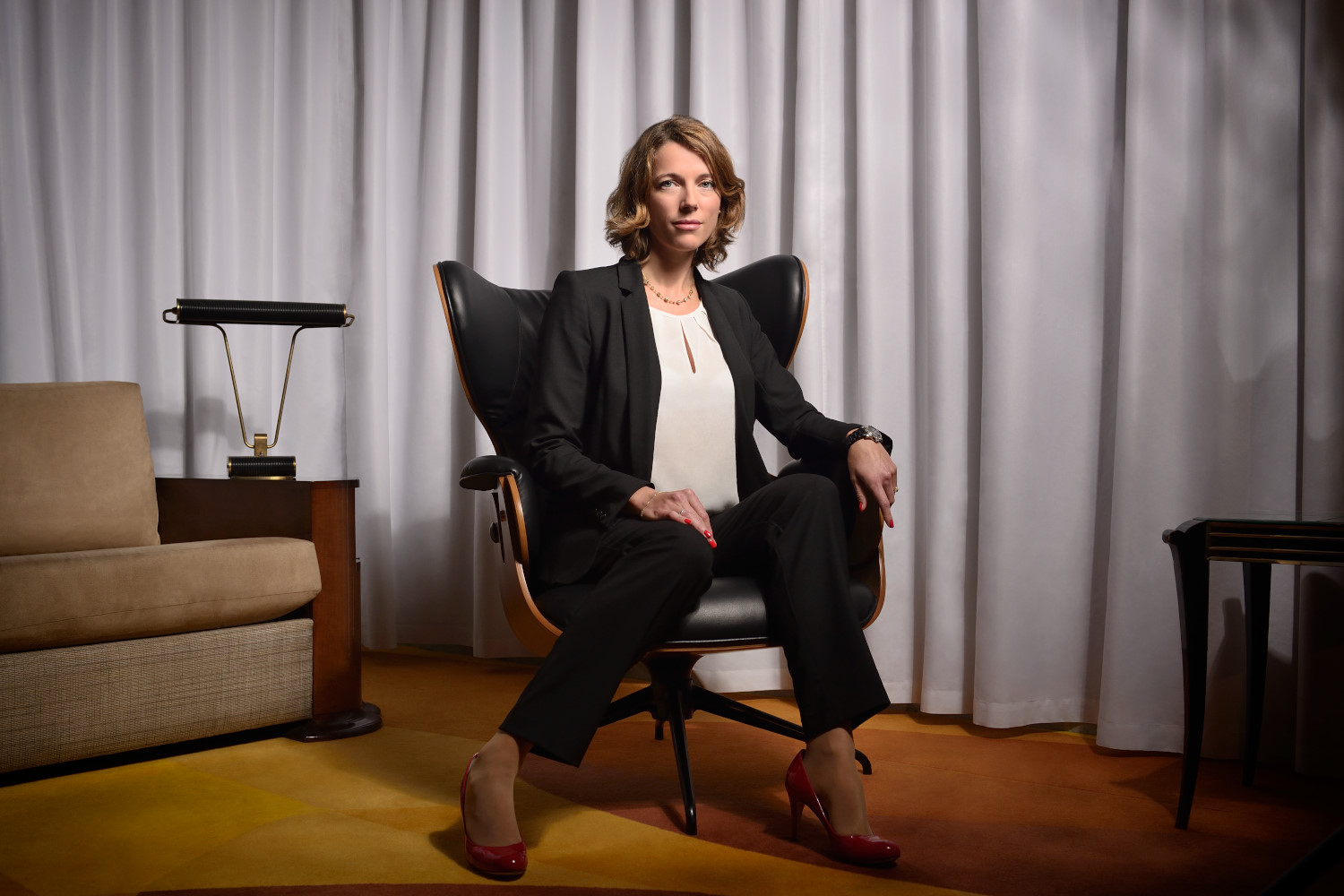 Une séance photo portrait pour CV LinkedIn et site web capture une femme en costume noir et talons rouges, assise avec assurance sur une chaise moderne devant des rideaux blancs. Avec des cheveux courts et ondulés et un regard direct vers la caméra, le décor sophistiqué comprend une table d'appoint et une lampe.