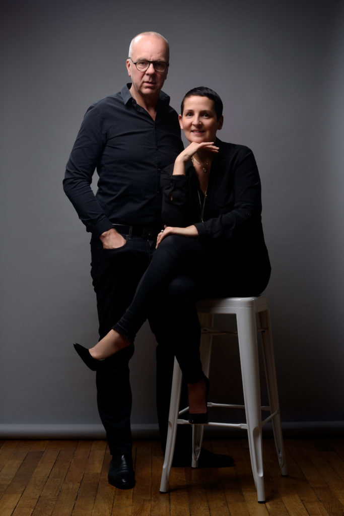 Un homme et une femme vêtus de noir posent devant un fond gris. L'homme se tient légèrement en retrait et à gauche avec une posture détendue, tandis que la femme est assise sur un grand tabouret, posant son menton sur sa main et croisant les jambes. Tous deux regardent directement vers l'appareil photo, créant une séance de portrait idéale pour les profils LinkedIn ou les sites Web.