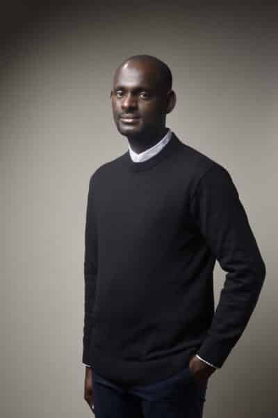 Un homme au crâne rasé, vêtu d'un pull noir sur une chemise à col blanc, pose sur un fond gris uni. Une main est dans sa poche, et il a une expression calme, parfaite pour une séance photo portrait pour CV LinkedIn et site web.