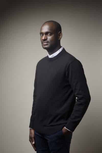 Un homme aux cheveux courts se tient debout sur un fond de couleur neutre, parfait pour une séance photo portrait pour CV LinkedIn et site web. Il porte un pull noir sur une chemise à col blanc, avec sa main gauche dans sa poche. Il a une expression calme et posée, regardant légèrement vers sa gauche.