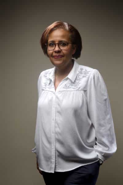 Une femme aux cheveux bruns courts et aux lunettes se tient debout sur un fond uni. Elle porte une blouse blanche avec des broderies florales sur les épaules et un pantalon foncé, reflétant le professionnalisme. Avec un sourire doux et les mains dans les poches, elle est parfaite pour une séance photo portrait pour CV LinkedIn et site web.