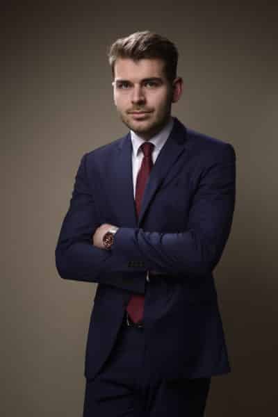 Un homme aux cheveux courts, vêtu d'un costume bleu marine, d'une chemise blanche et d'une cravate rouge, se tient debout, les bras croisés, sur un fond uni marron clair. Il porte également une montre-bracelet au poignet gauche et a une expression neutre, ce qui le rend idéal pour une séance photo portrait pour CV LinkedIn et site web.