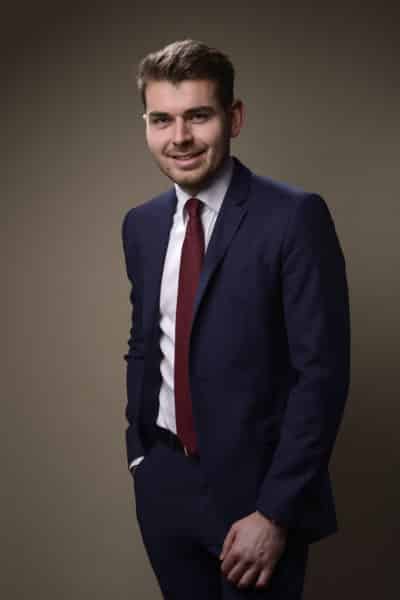 Un homme vêtu d'un costume bleu foncé, d'une chemise blanche et d'une cravate rouge se tient debout sur un fond uni et sombre. Il sourit avec une main dans sa poche pendant une séance photo portrait pour CV LinkedIn et site web.