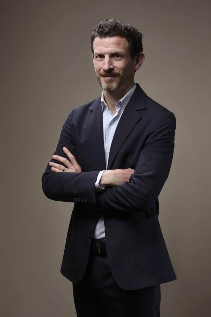 Une séance photo portrait pour CV LinkedIn et site web capture une personne avec des cheveux courts et une barbe taillée, debout contre un fond universitaire. Vêtu d'une veste de costume sombre et d'une chemise bleu clair, les bras croisés, il regarde la caméra avec assurance.