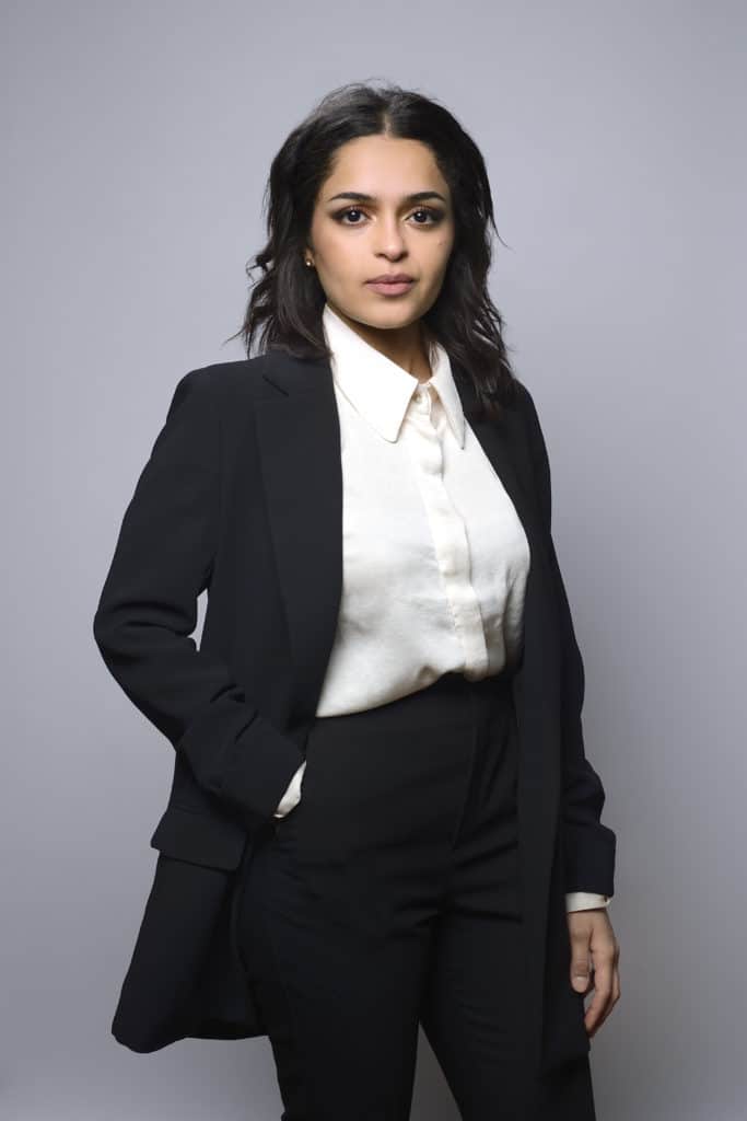 Une femme aux cheveux noirs mi-longs, vêtue d'un chemisier blanc et d'un blazer noir, se tient debout sur un fond gris uni. Une main est dans sa poche tandis qu'elle regarde avec assurance l'appareil photo, capturant parfaitement l'essence d'une séance photo portrait pour CV LinkedIn et site web.