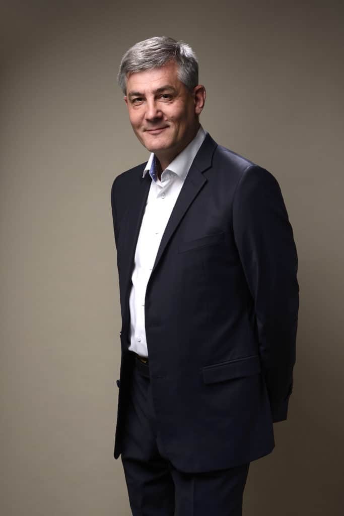 Un homme aux cheveux gris se tient debout sur un fond uni, vêtu d'un costume sombre et d'une chemise blanche. Il a les mains derrière le dos et sourit légèrement à la caméra pendant une séance photo portrait pour CV LinkedIn et site web.