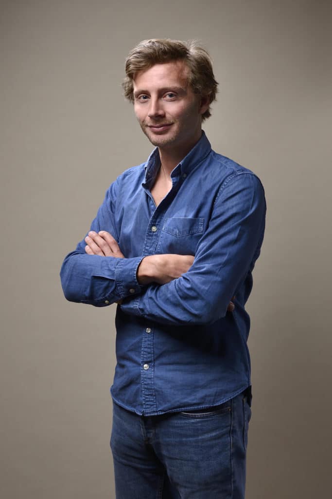 Un homme aux cheveux blonds courts se tient debout sur un fond uni, idéal pour un portrait photo de séance pour CV linkedin et site web. Il porte une chemise bleue boutonnée et un jean, les bras croisés et un léger sourire sur le visage.