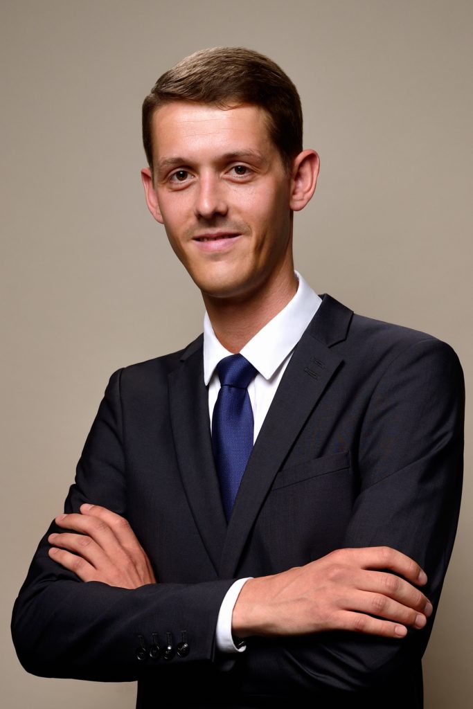Un jeune homme aux cheveux bruns courts, vêtu d'un costume sombre, d'une chemise blanche et d'une cravate bleu foncé, se tient debout, les bras croisés, devant un fond neutre. Il regarde la caméra avec un léger sourire, parfait pour une séance photo portrait pour CV LinkedIn et site web.
