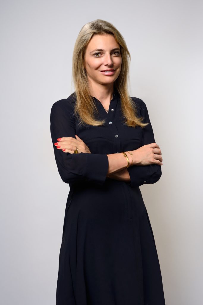 Une femme aux longs cheveux blonds se tient debout sur un fond uni, vêtue d'une robe noire boutonnée. Elle sourit, les bras croisés, avec du vernis à ongles rouge et un bracelet sur une main. Ce portrait photo d'une séance pour CV LinkedIn et site web capture son attitude professionnelle mais accessible.