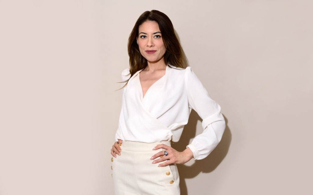 Une femme aux longs cheveux noirs se tient debout, les mains sur les hanches, avec assurance. Elle porte un chemisier blanc élégant à col en V et une jupe taille haute assortie ornée de boutons dorés, prête pour une séance photo portrait pour CV LinkedIn et site web. L'arrière-plan est d'une couleur unie et claire.